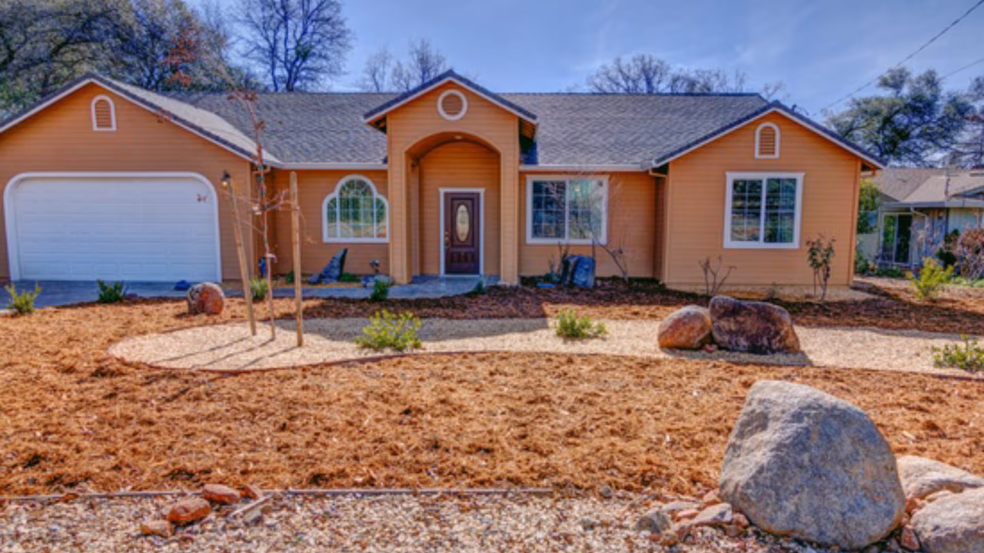 Front exterior view of a single-story house with an orange facade, a dark brown front door with an oval glass panel, and a two-car garage. The yard features mulch, rocks, small plants, and a few young trees under a clear blue sky.