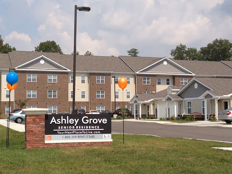 Exterior view of Ashley Grove Senior Residence, a multi-story senior living apartment building with a brick and beige facade. There is a large sign in front displaying the facility name, website, and phone number. Balloons are tied to poles near the sign, and several cars are parked in the driveway and parking area. Trees and a partly cloudy sky are visible in the background.