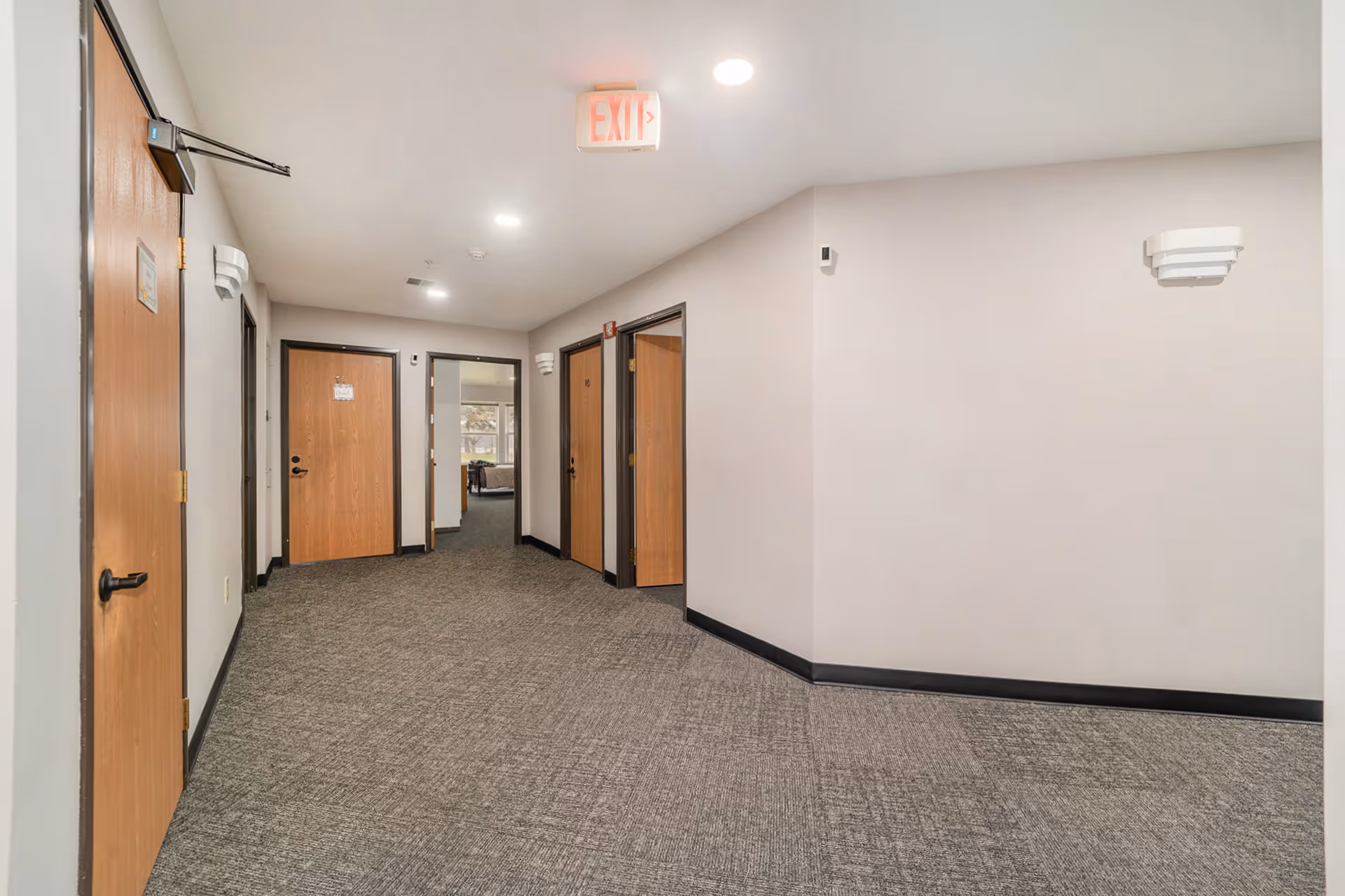Interior hallway of Pine Tree Place senior living facility with multiple wooden doors, gray carpeted floor, white walls, ceiling lights, and an exit sign above one door. A room with a bed is visible at the end of the hallway.