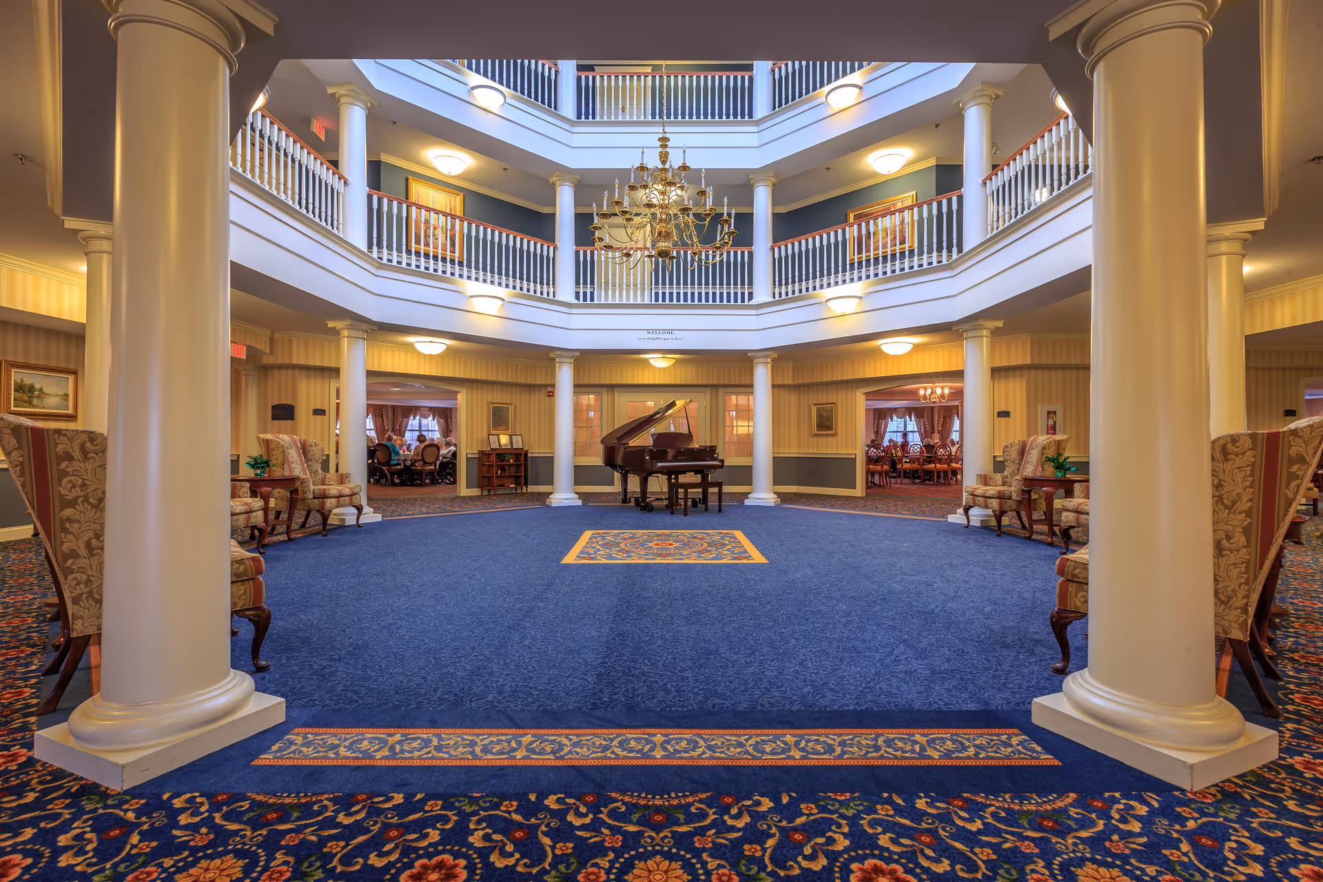Spacious and elegant interior common area with a grand piano in the center, surrounded by white columns and a blue carpet with intricate patterns. The area is well-lit with wall sconces and a large chandelier hanging from the ceiling. There are seating areas with upholstered chairs along the sides and a view of dining rooms in the background.