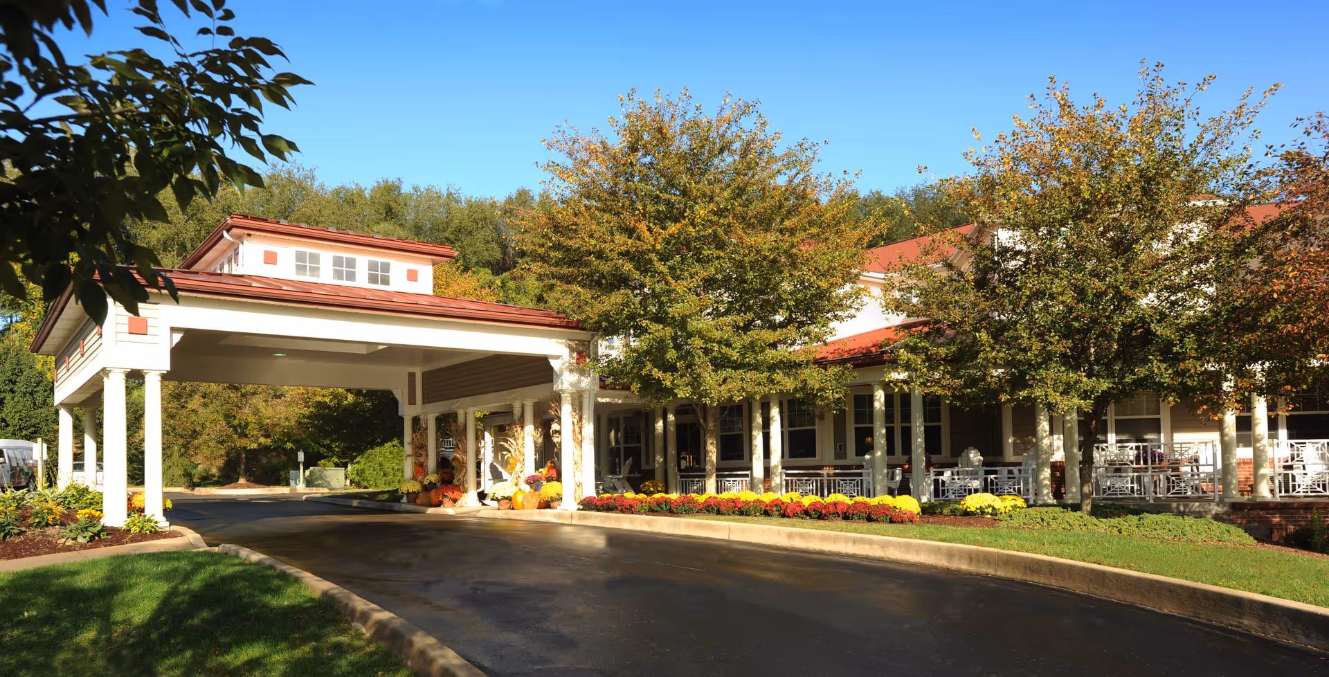 Front entrance and porte-cochere of a senior living facility with a curved driveway, landscaped flower beds, and trees.