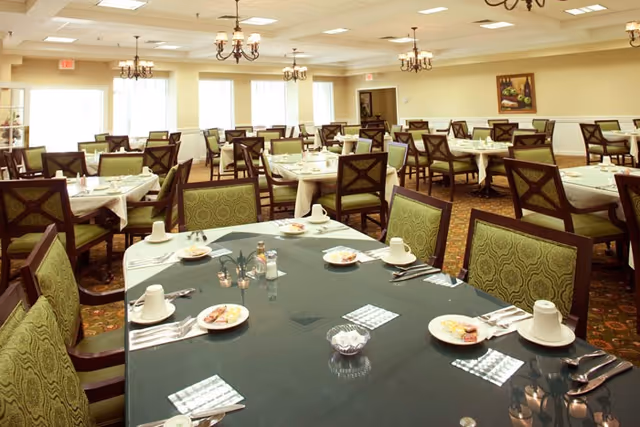 Spacious dining room with multiple tables set for meals, green upholstered chairs, and chandeliers.