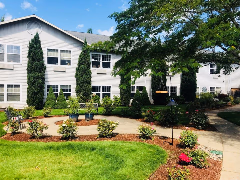 Outdoor garden area at Tanner Spring Assisted Living & Memory Care featuring a well-maintained lawn, flower beds with blooming roses, potted plants, a concrete pathway, benches, and tall evergreen trees in front of a two-story white building under a blue sky with some clouds.