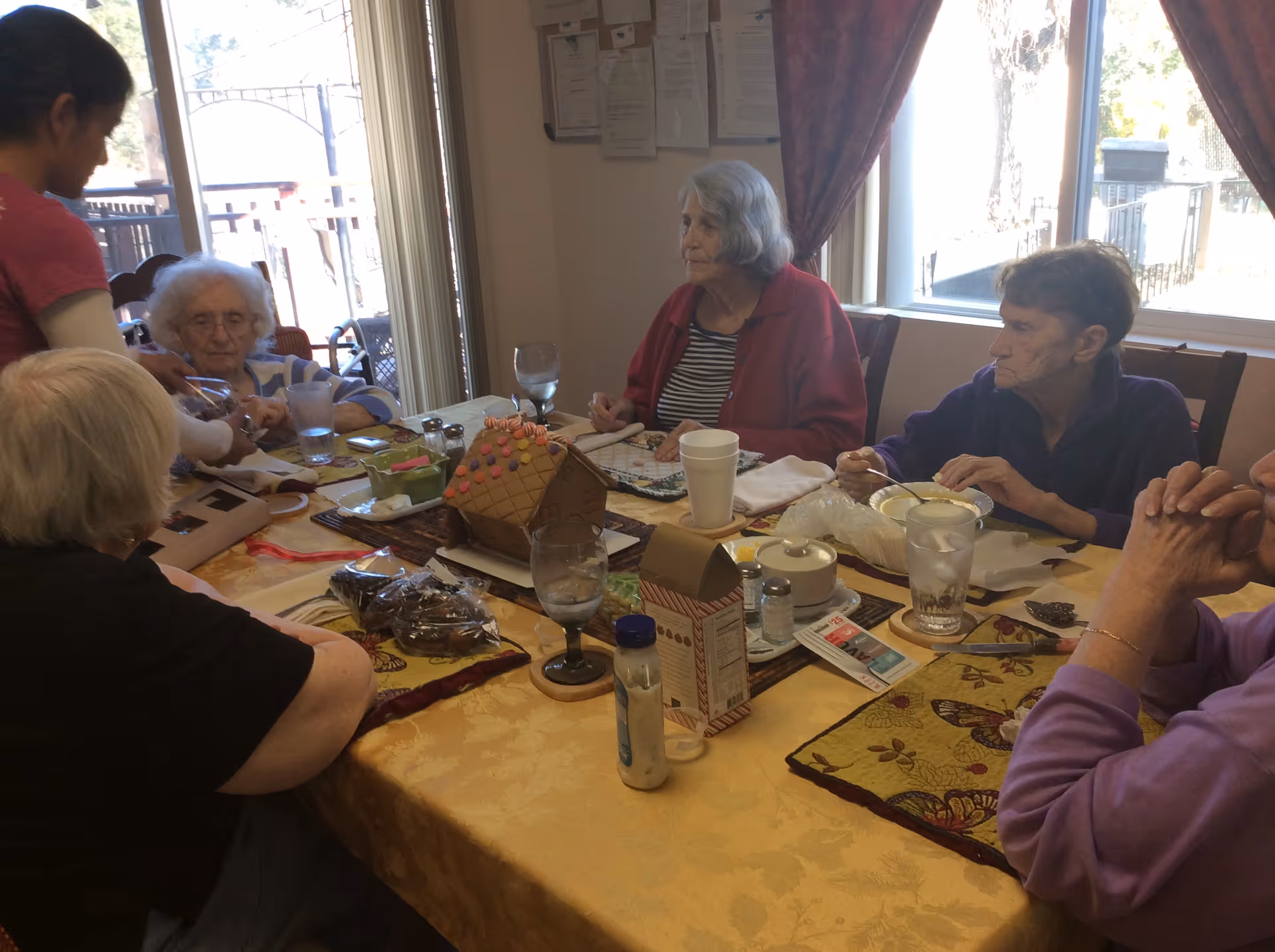 A group of elderly women sit around a dining table with plates, glasses, and a decorated gingerbread house in a communal dining area.