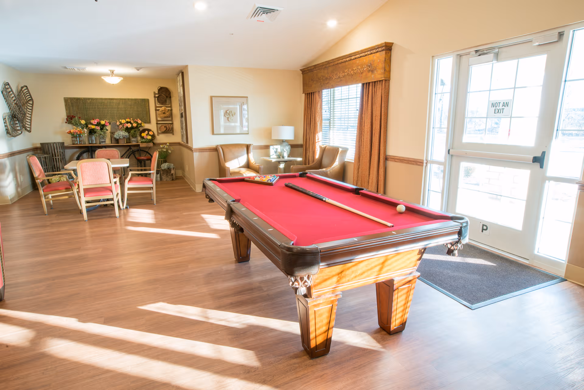 A bright room with a red felt pool table in the center, a cue stick and billiard balls on the table. To the left, there is a small dining area with a table and chairs, and a wall decorated with flowers and artwork. Two armchairs and a side table with a lamp are positioned near a window with brown curtains. A glass door with a 'Not An Exit' sign is visible on the right side.