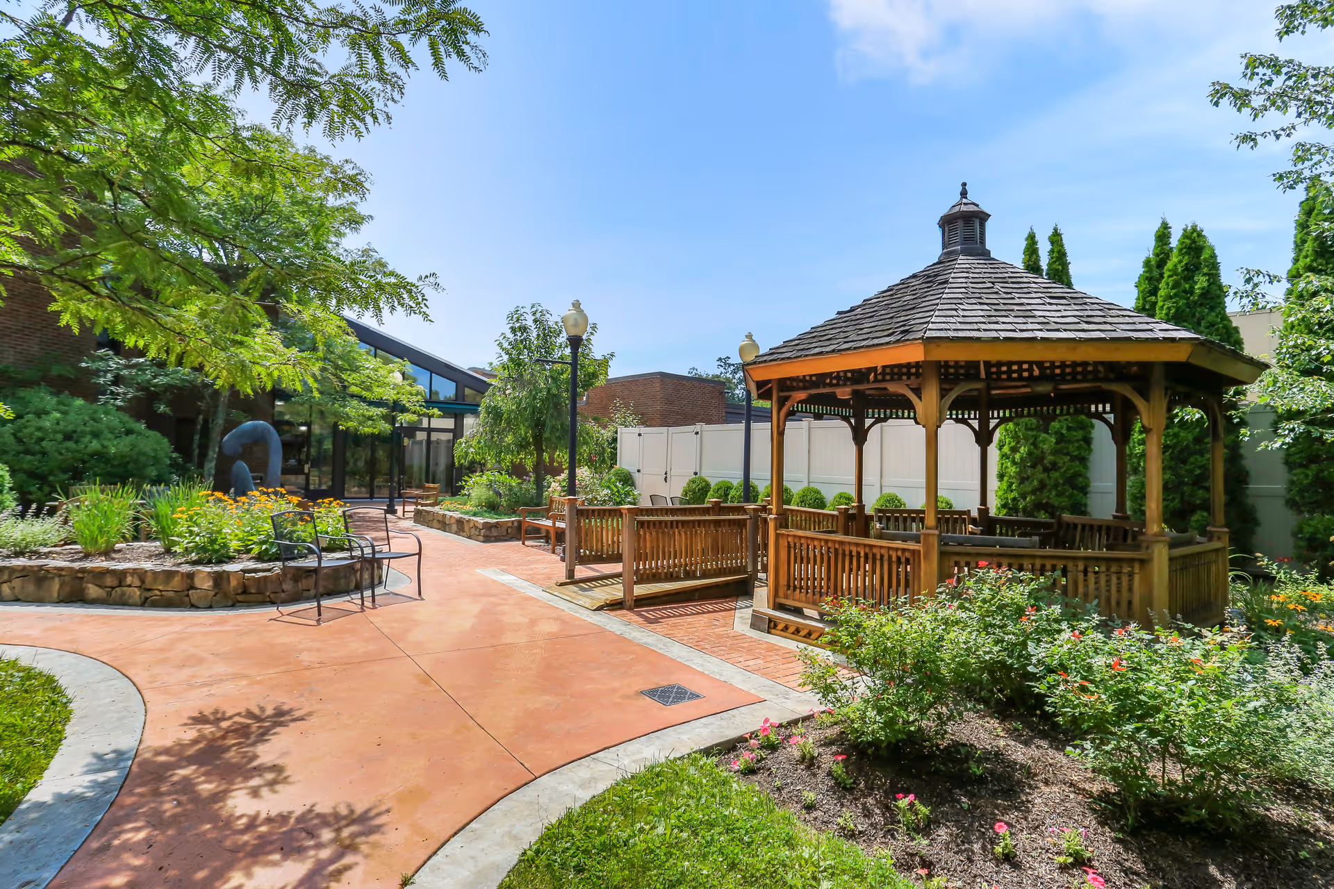 Outdoor garden area at The Village at Waveny featuring a wooden gazebo with benches, surrounded by green trees, shrubs, and colorful flowers under a clear blue sky. There are paved walkways and a few chairs near a raised flower bed made of stone.