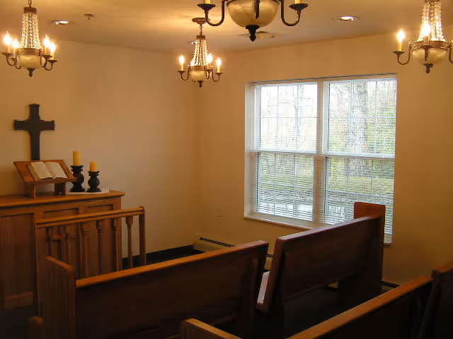Small chapel room with wooden pews facing a wooden altar that holds an open Bible, two candles, and a cross mounted on the wall. The room is lit by multiple chandeliers and has a window with blinds showing trees outside.