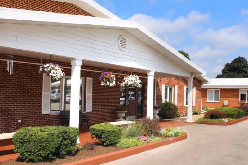Exterior view of a brick building with white columns supporting a covered entrance. Hanging flower baskets with colorful blooms are suspended from the roof. There are neatly trimmed bushes and flower beds along the walkway leading to the entrance. The sky is blue with some clouds.