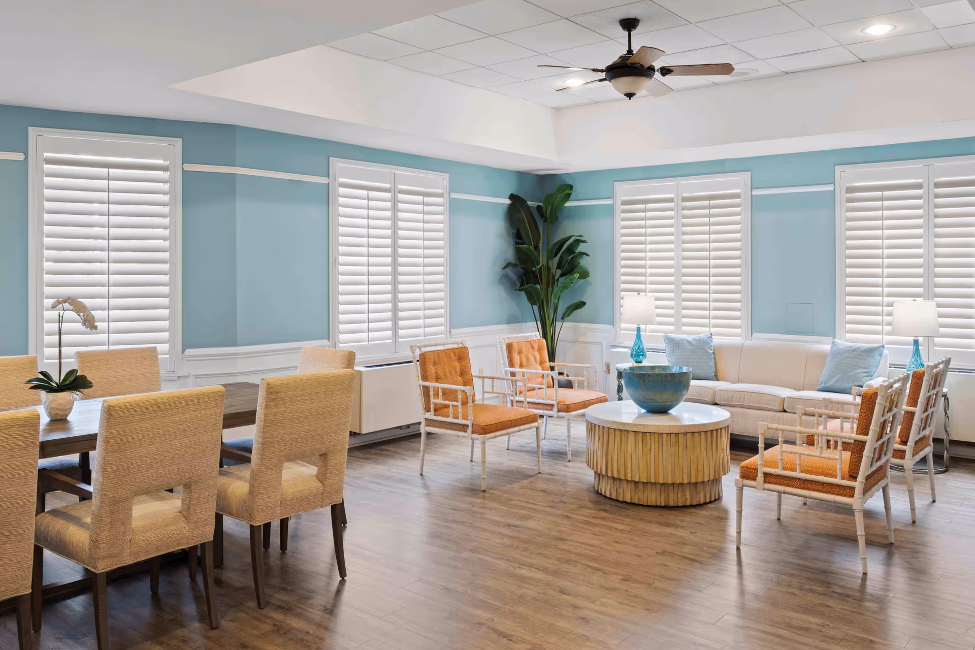 A bright and spacious living room area with light blue walls and white window shutters. The room features a beige sofa with light blue pillows, four orange cushioned chairs with white frames arranged around a round coffee table with a decorative bowl. To the left, there is a wooden dining table with six beige upholstered chairs and a small potted plant on top. The floor is wood, and there is a ceiling fan with lights above.