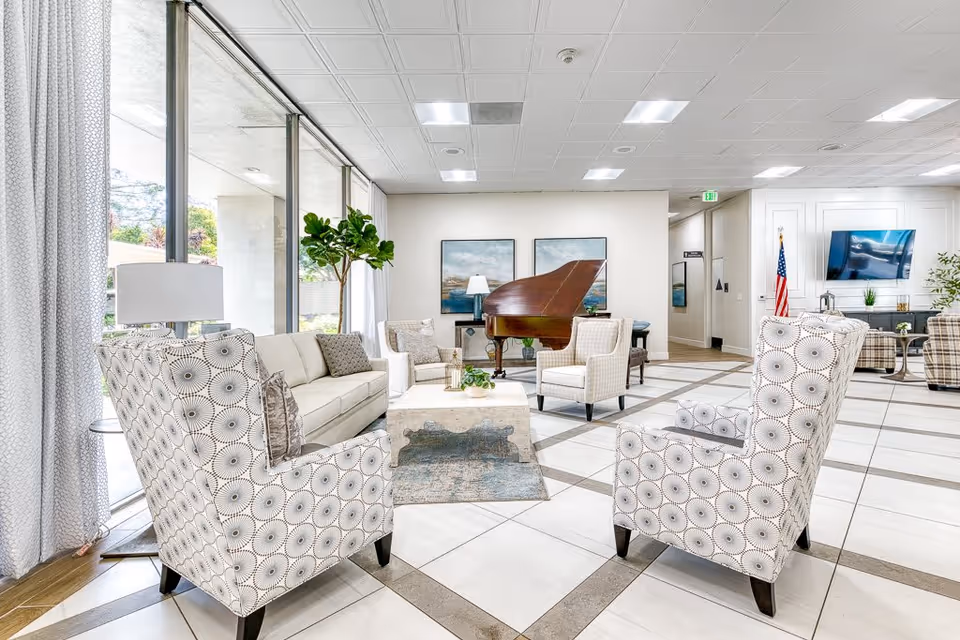 A bright and spacious senior living facility common area with patterned armchairs, a beige sofa, a coffee table, a grand piano, and large windows letting in natural light. The room features tiled floors, white walls, framed artwork, and an American flag near a wall-mounted TV.