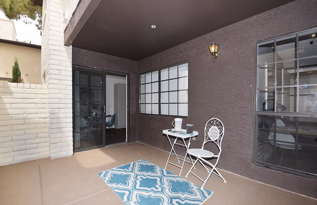A covered outdoor patio area with a small white metal table and matching chair on a brown floor. A blue and white patterned rug is placed in front of the table. The patio has dark brown walls with a window and a sliding glass door leading inside. A wall-mounted light fixture is above the table. The adjacent area outside the patio has a white brick wall and some greenery visible.