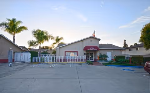 Exterior view of a single-story building with a red awning over the entrance, palm trees around the property, and a parking lot in front with marked parking spaces including a handicapped spot.