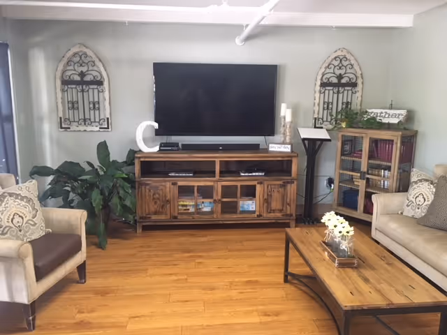 A cozy living room with wooden flooring featuring a flat-screen TV mounted on a light gray wall above a wooden TV stand. The room includes a beige armchair with patterned cushions on the left, a beige sofa with cushions on the right, and a wooden coffee table with a flower arrangement in the center. Decorative wall hangings and a green plant add to the room's inviting atmosphere.