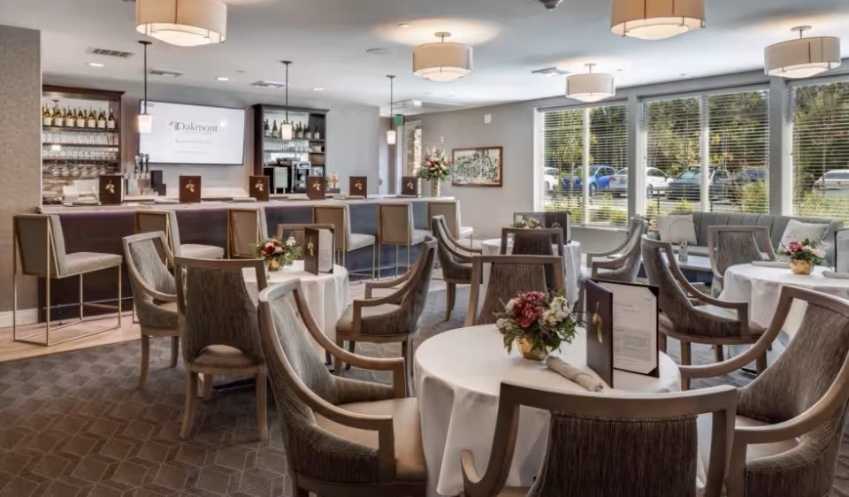 A well-lit dining area with round tables covered in white tablecloths, each adorned with floral centerpieces and menus. The room features comfortable chairs around the tables and a bar area with high stools in the background. Large windows with blinds allow natural light to fill the space, and pendant lights hang from the ceiling.