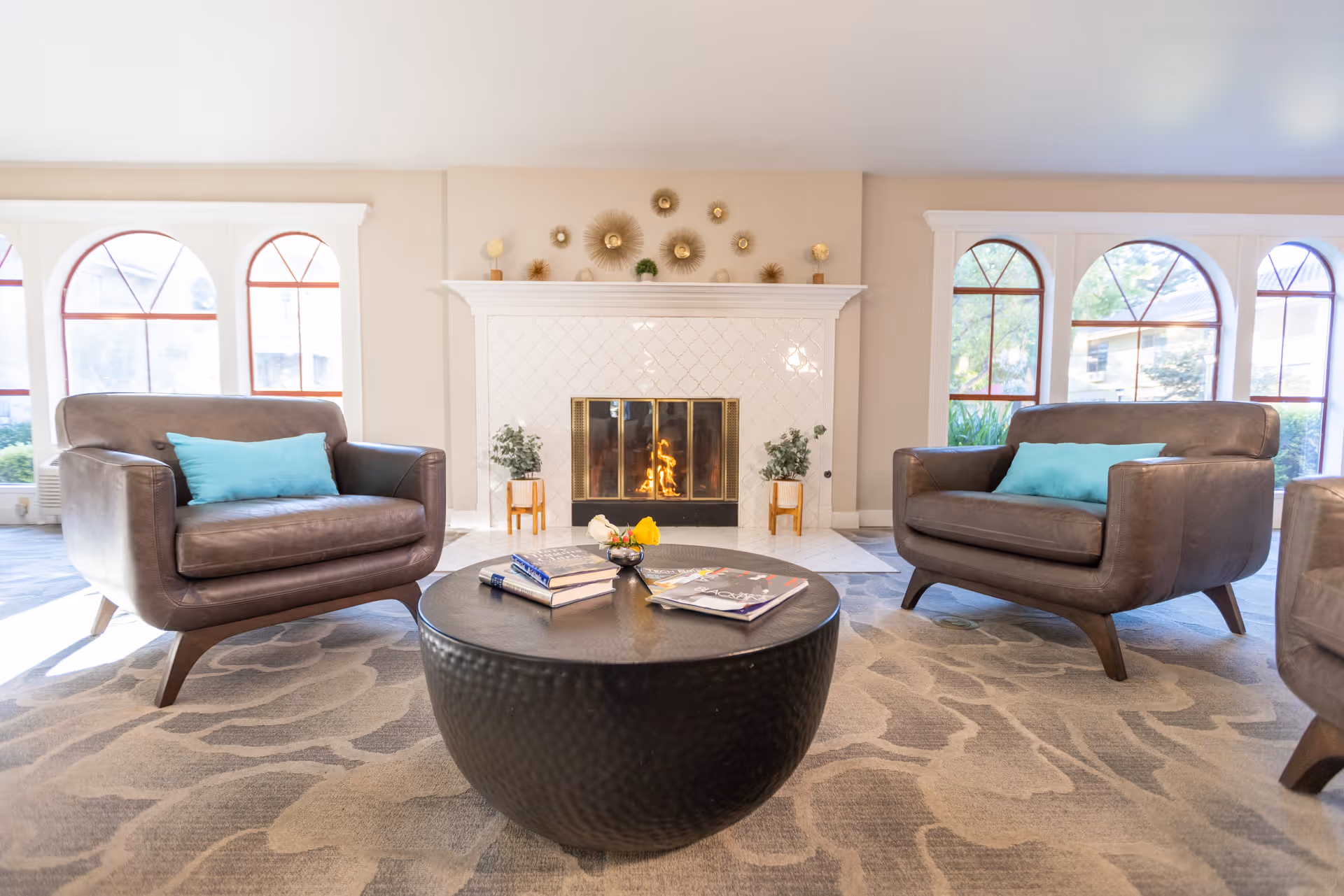 A cozy living room area with two brown leather armchairs each with a blue cushion, a round black coffee table with books and a small decorative item, a lit fireplace with a white tiled surround, and large arched windows letting in natural light.