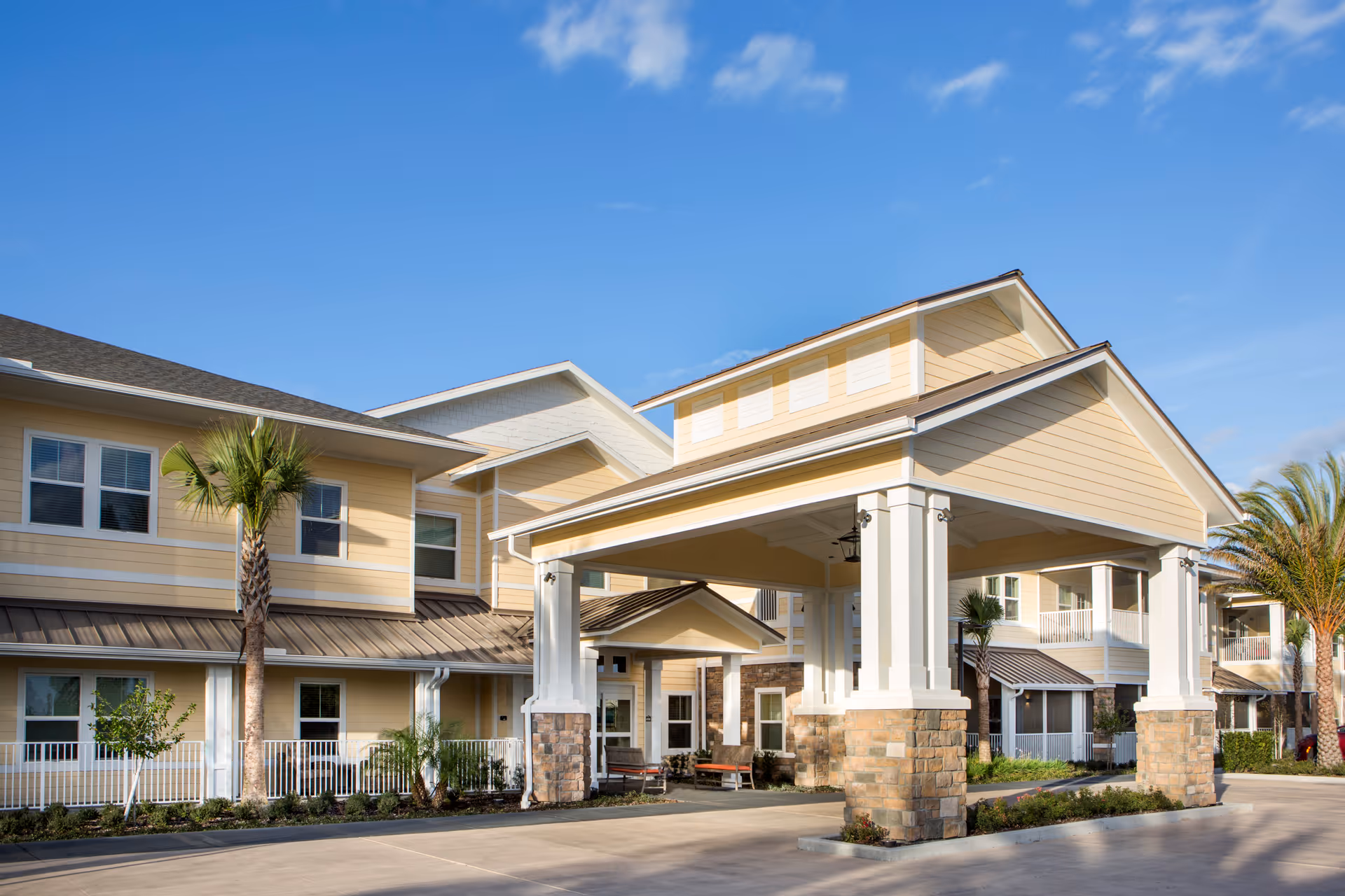 Exterior view of The Summit of Winter Garden senior living facility showing a covered entrance with stone pillars, beige siding, multiple windows, palm trees, and a clear blue sky.