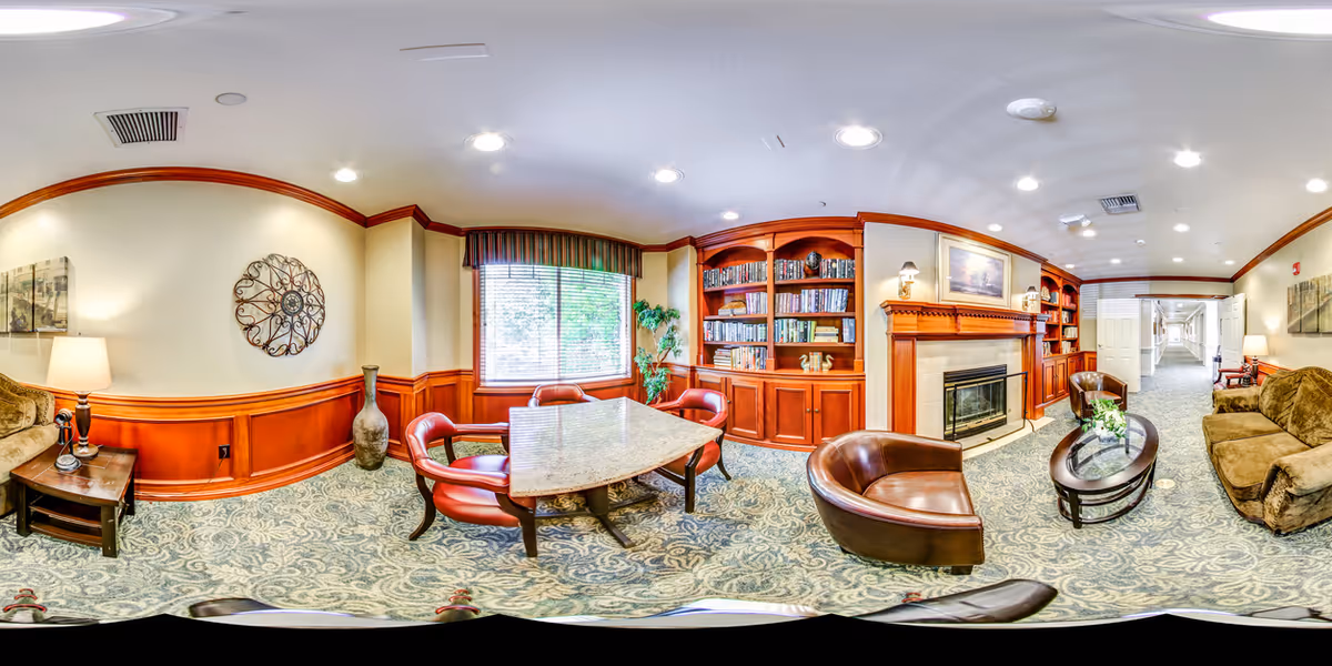 A cozy living room area in a senior living facility featuring a fireplace with a wooden mantel, built-in bookshelves filled with books, a large window with blinds and a valance, a square table with four red cushioned chairs, two brown leather armchairs, a brown sofa, a wooden coffee table with a small plant, a side table with a lamp, and decorative wall art. The room has carpeted flooring with a patterned design and wood paneling on the lower half of the walls.