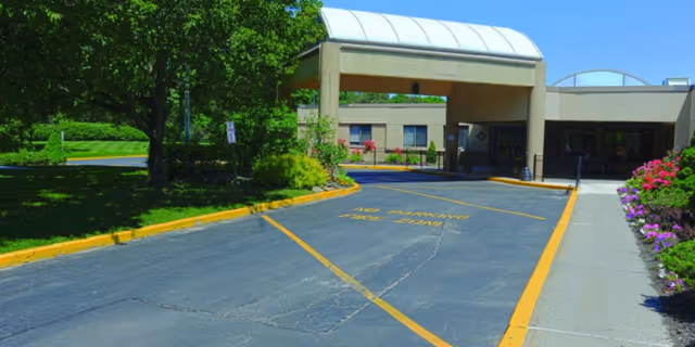 Entrance driveway to a healthcare facility with a covered drop-off area, surrounded by greenery and flowers on a sunny day.