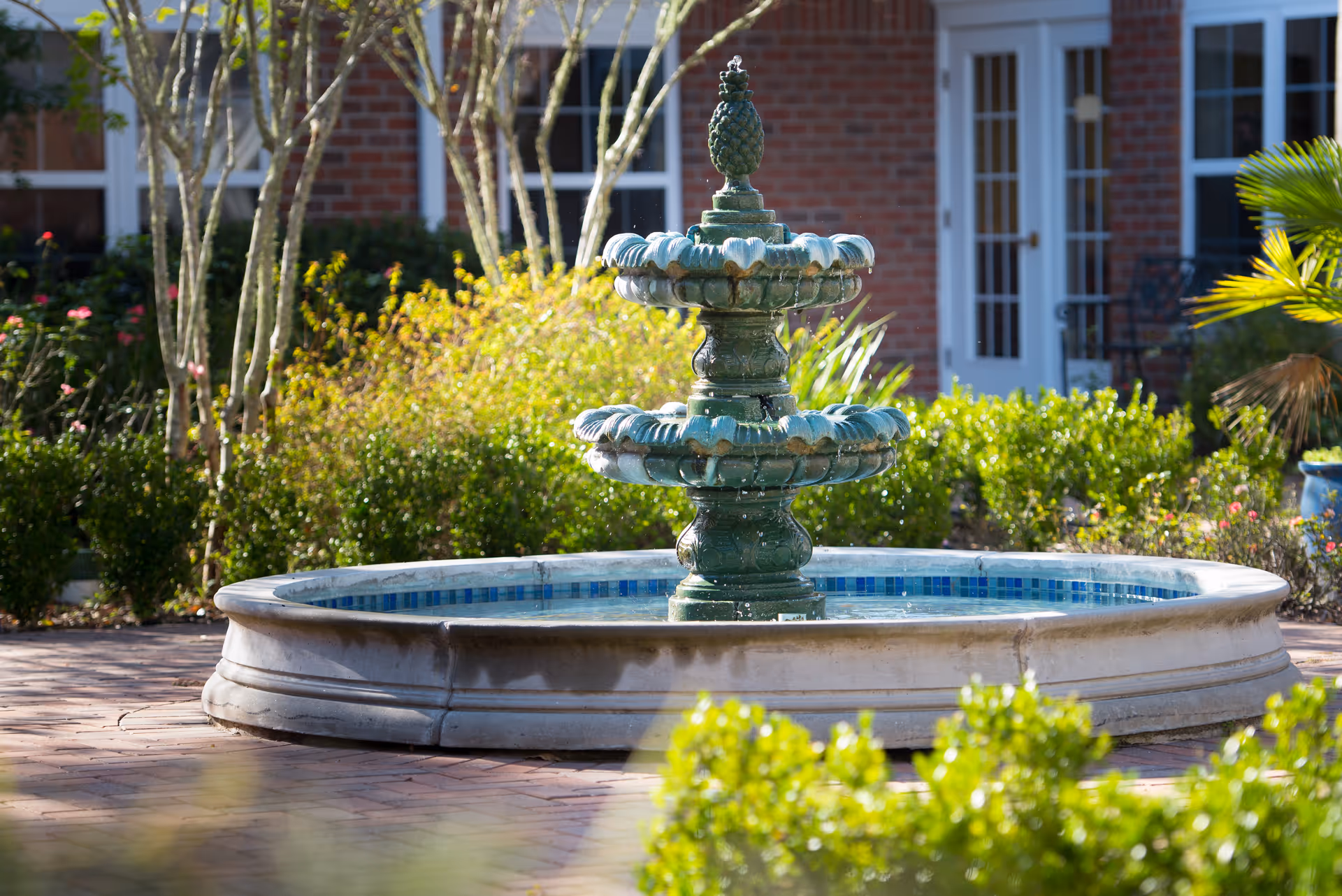 A decorative two-tier green fountain in a landscaped courtyard in front of a brick building.