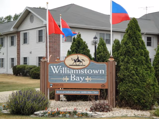 Exterior view of Williamstown Bay senior living facility with a large sign displaying the facility name, surrounded by landscaping including bushes and flowers, and three flagpoles with red and blue flags in front of a two-story building.