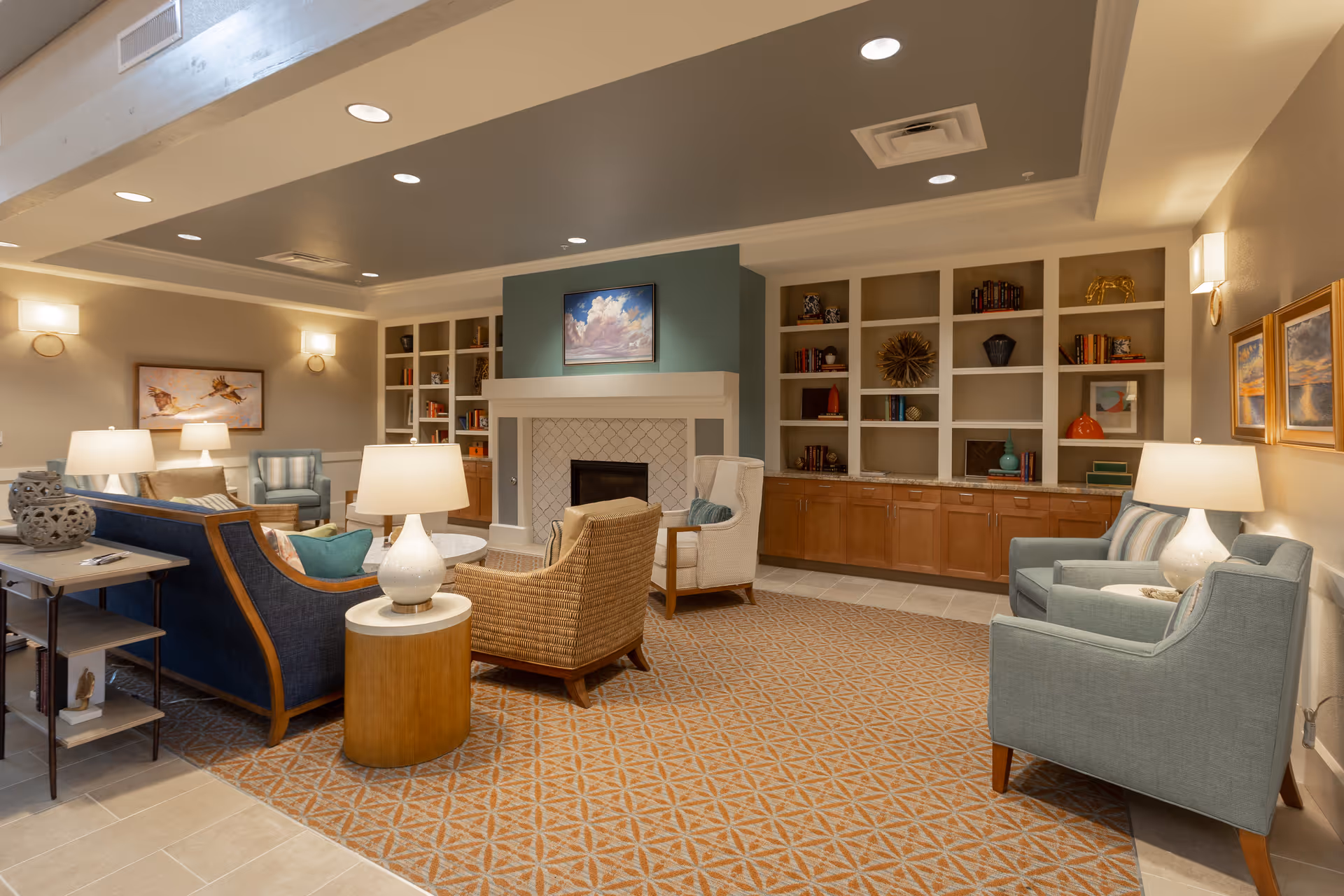 A cozy living room area in a senior living facility featuring a fireplace with a painting above it, built-in shelves filled with books and decorative items, multiple armchairs and sofas arranged around a central coffee table, warm lighting from table lamps and wall sconces, and a patterned carpet covering the floor.