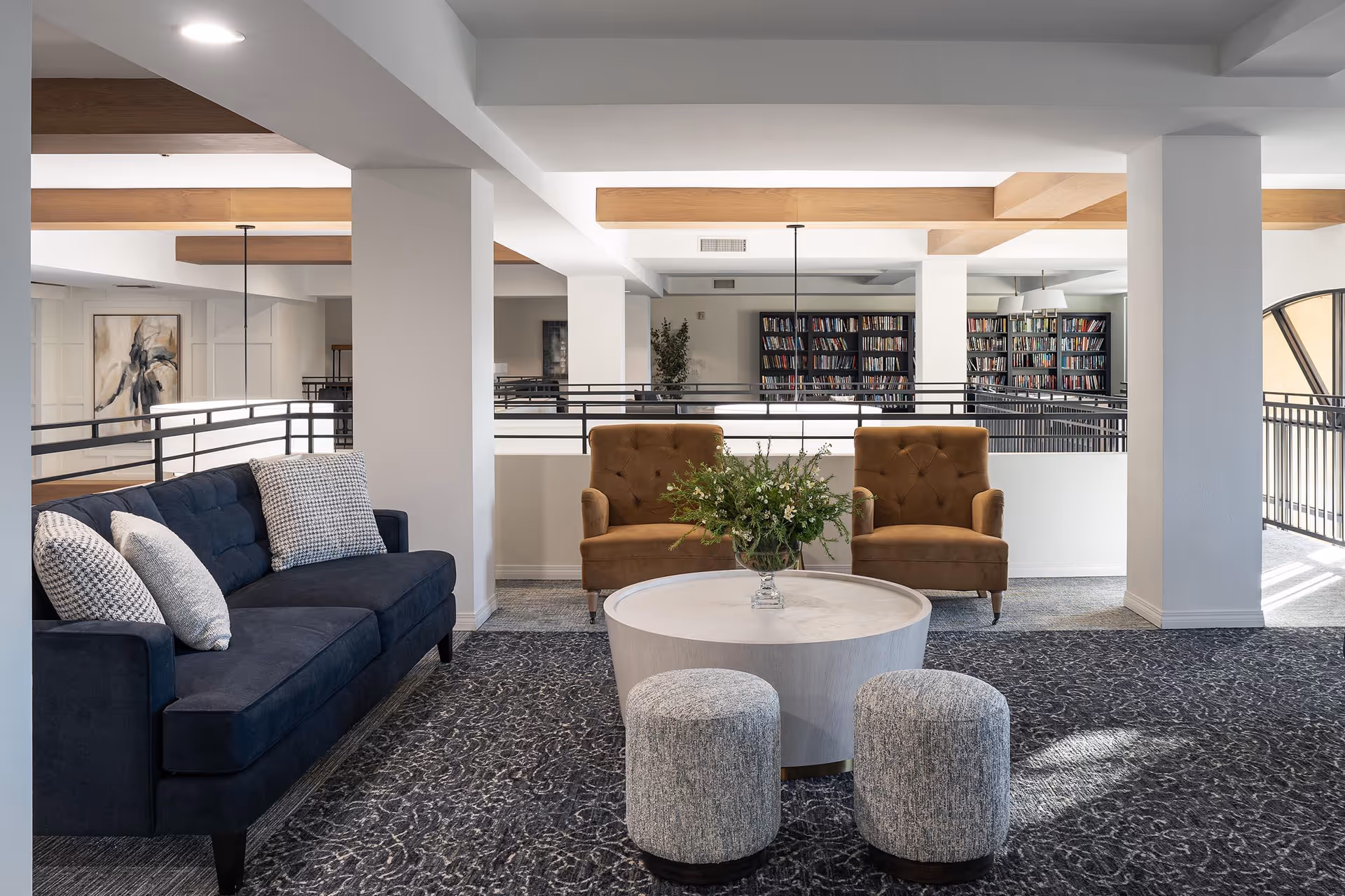 A modern and cozy living room area with a dark blue sofa adorned with patterned cushions on the left, two brown armchairs in the center back, and two small gray ottomans in front of a round white coffee table with a floral arrangement. The room features white walls, wooden ceiling beams, a patterned carpet, and bookshelves in the background.