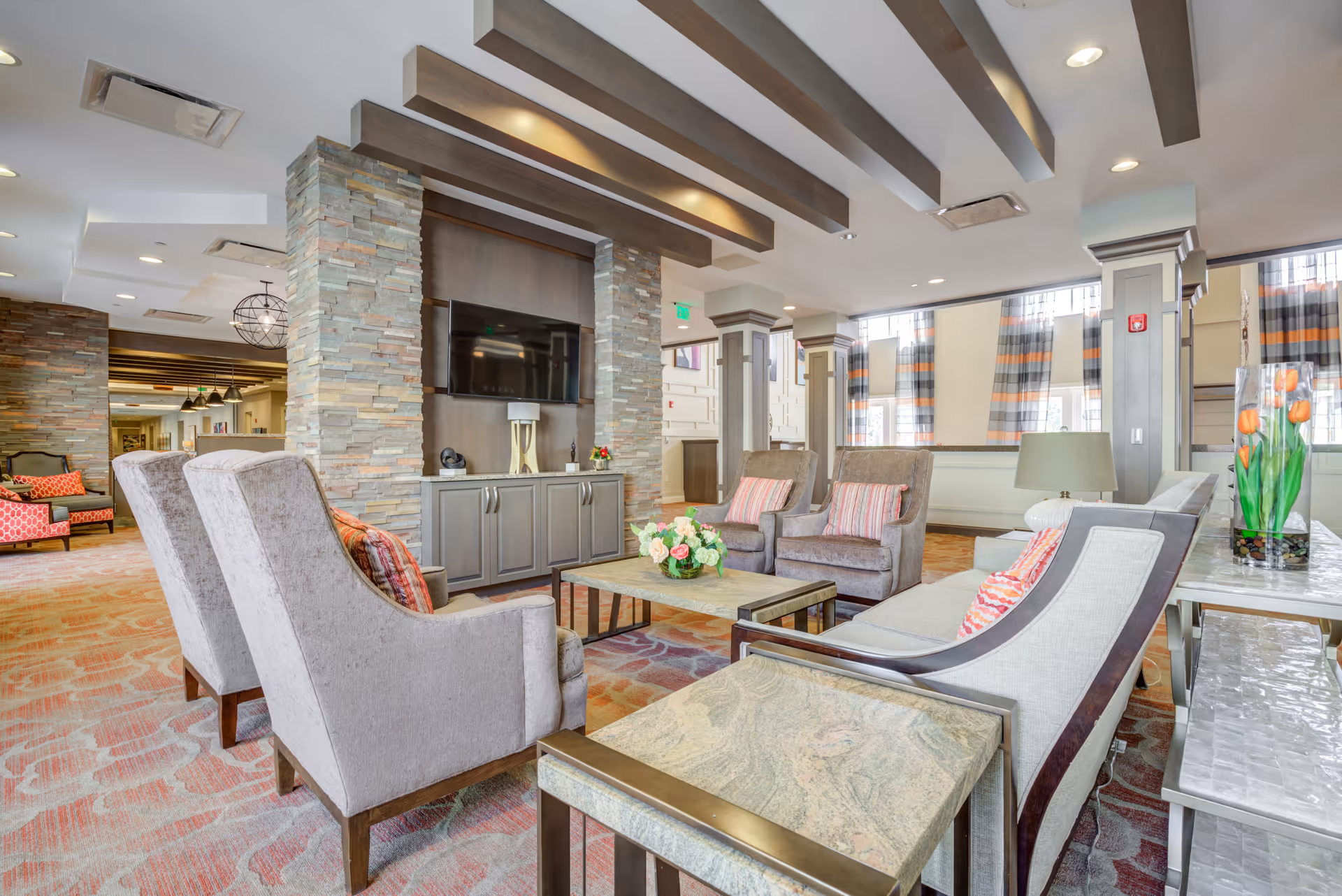 Spacious furnished communal lounge with armchairs, sofas, coffee tables, a wall-mounted TV between stone pillars, and decorative ceiling beams.