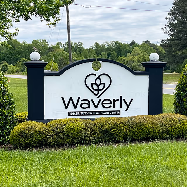 Outdoor sign for Waverly Rehabilitation and Healthcare Center surrounded by green grass and bushes, with trees and a road in the background under a cloudy sky.