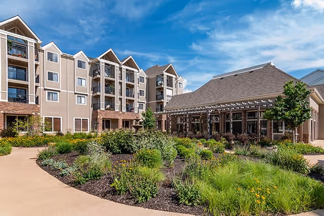 Landscaped courtyard and garden in front of a multi-story senior living building with balconies and a covered patio under a blue sky.