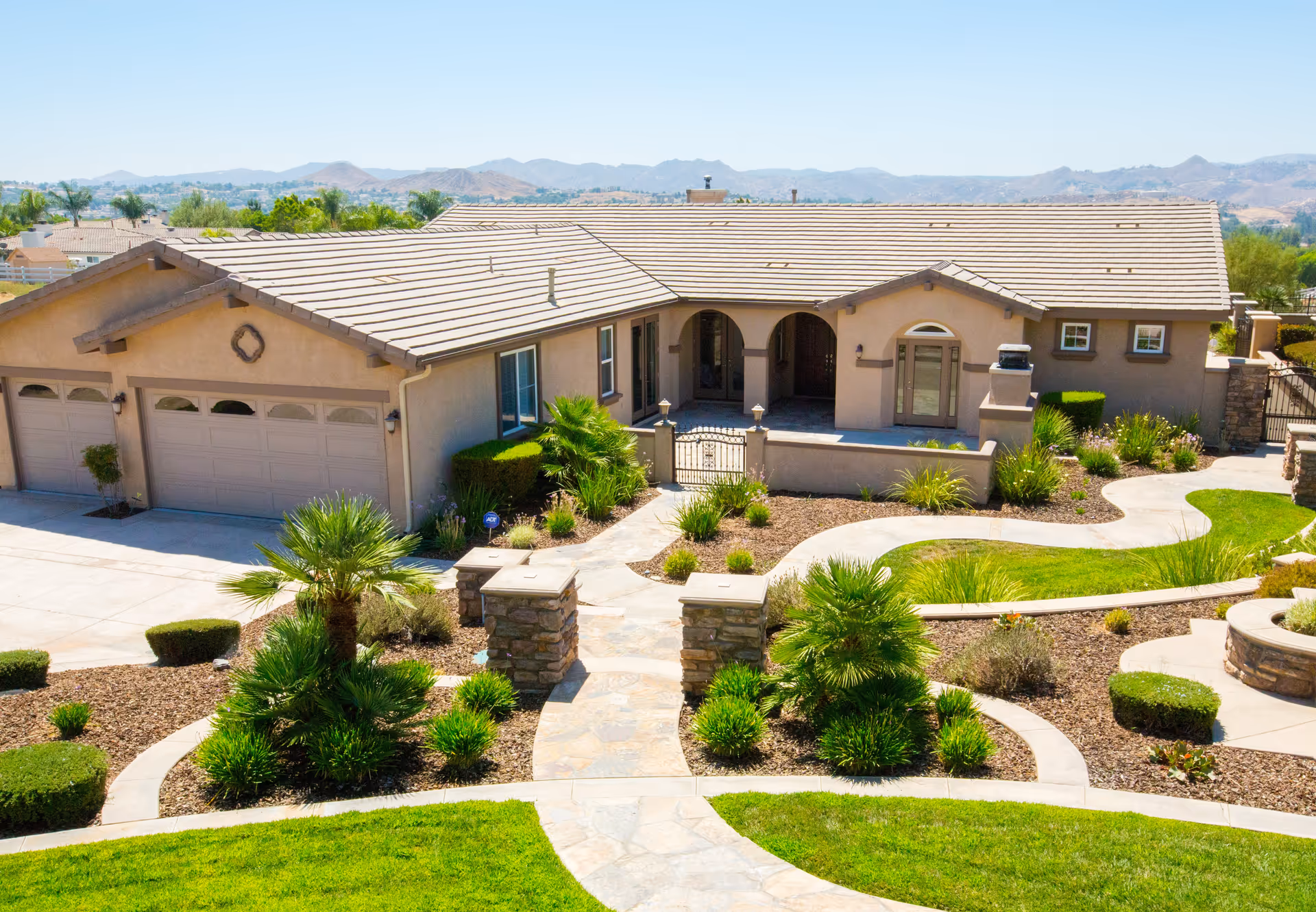 Front exterior view of a single-story senior living facility building with a tiled roof, a three-car garage, arched entryway, and landscaped garden with palm trees and shrubs under a clear blue sky.