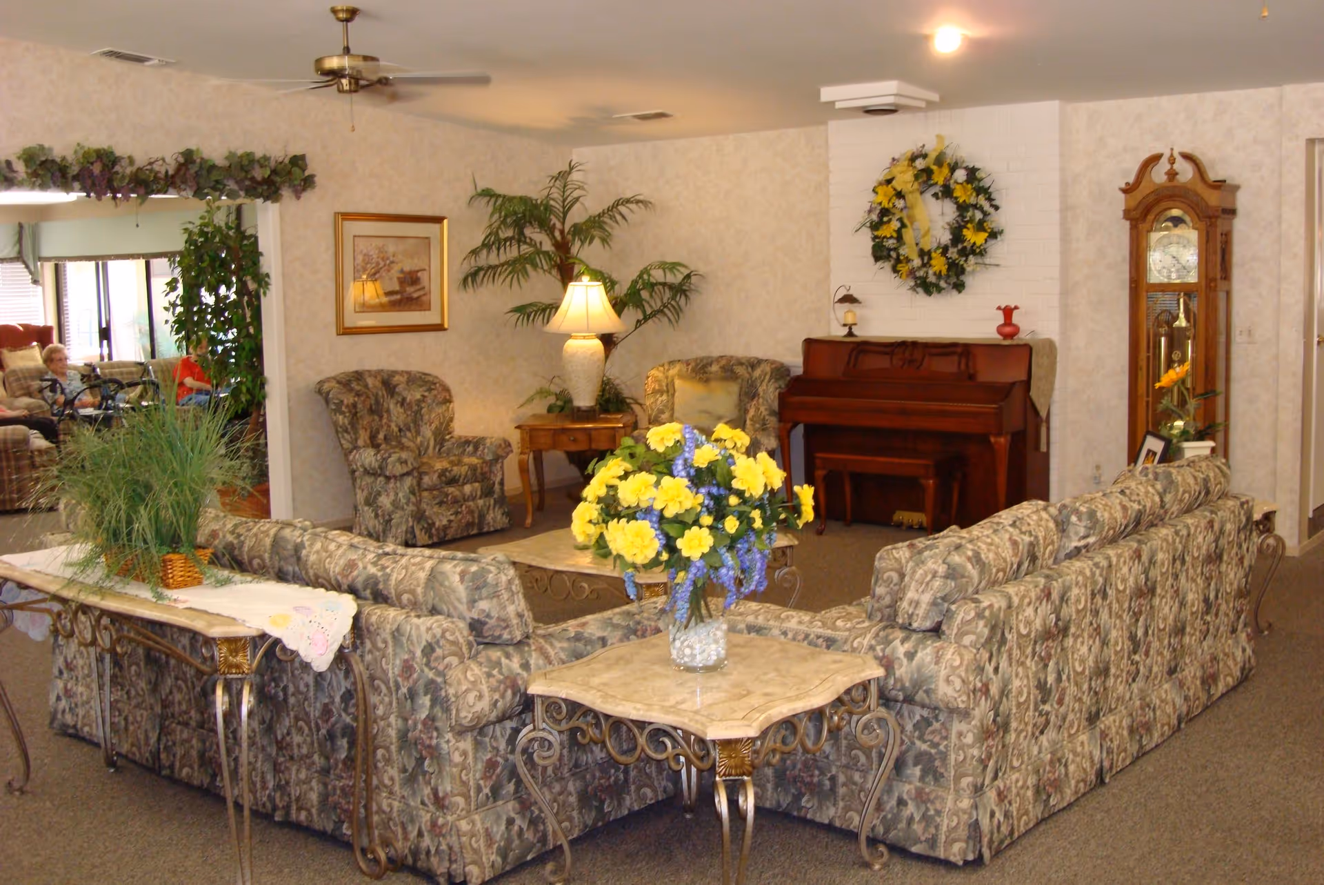 A cozy living room area in a senior living facility with floral patterned sofas arranged around a coffee table. There is a piano against the wall decorated with a yellow and white floral wreath above it. A grandfather clock stands to the right of the piano. The room has a ceiling fan, table lamps, framed artwork, and various plants including a vase of yellow and purple flowers on a side table.