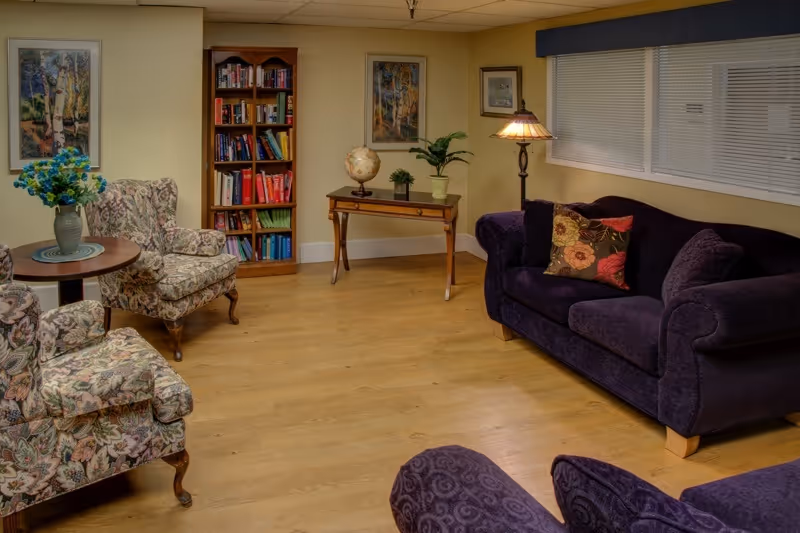 A cozy living room with wooden flooring, featuring a dark purple sofa with floral cushions, two patterned armchairs, a small round table with a vase of blue flowers, a wooden bookshelf filled with books, a wooden console table with a globe and plants, and framed artwork on the walls.