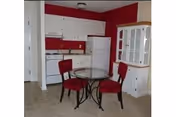 Small kitchen area with white cabinets, a white refrigerator, and a white hutch against a red accent wall. In front of the kitchen is a round glass-top table with a black metal base and three red cushioned chairs.