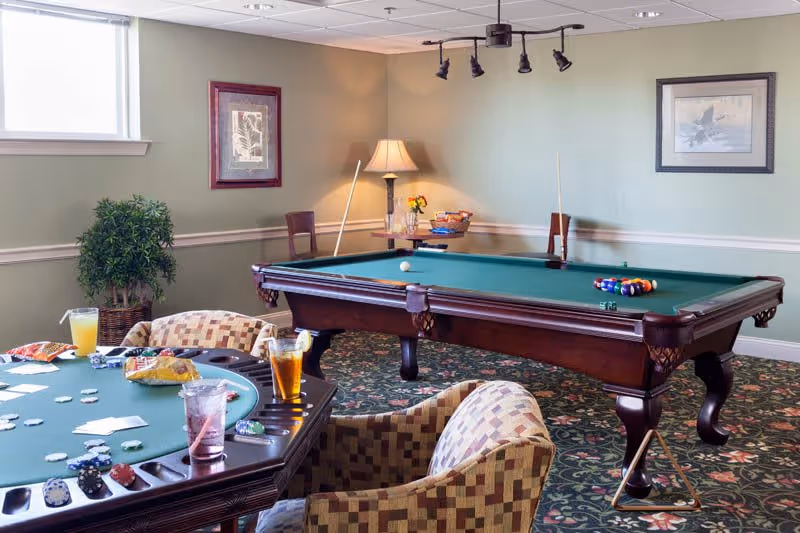 A recreational room with a pool table set up for a game and a poker table with playing cards, poker chips, snacks, and drinks. The room has patterned carpet, two framed pictures on the walls, a potted plant, and a lamp on a small table in the corner.