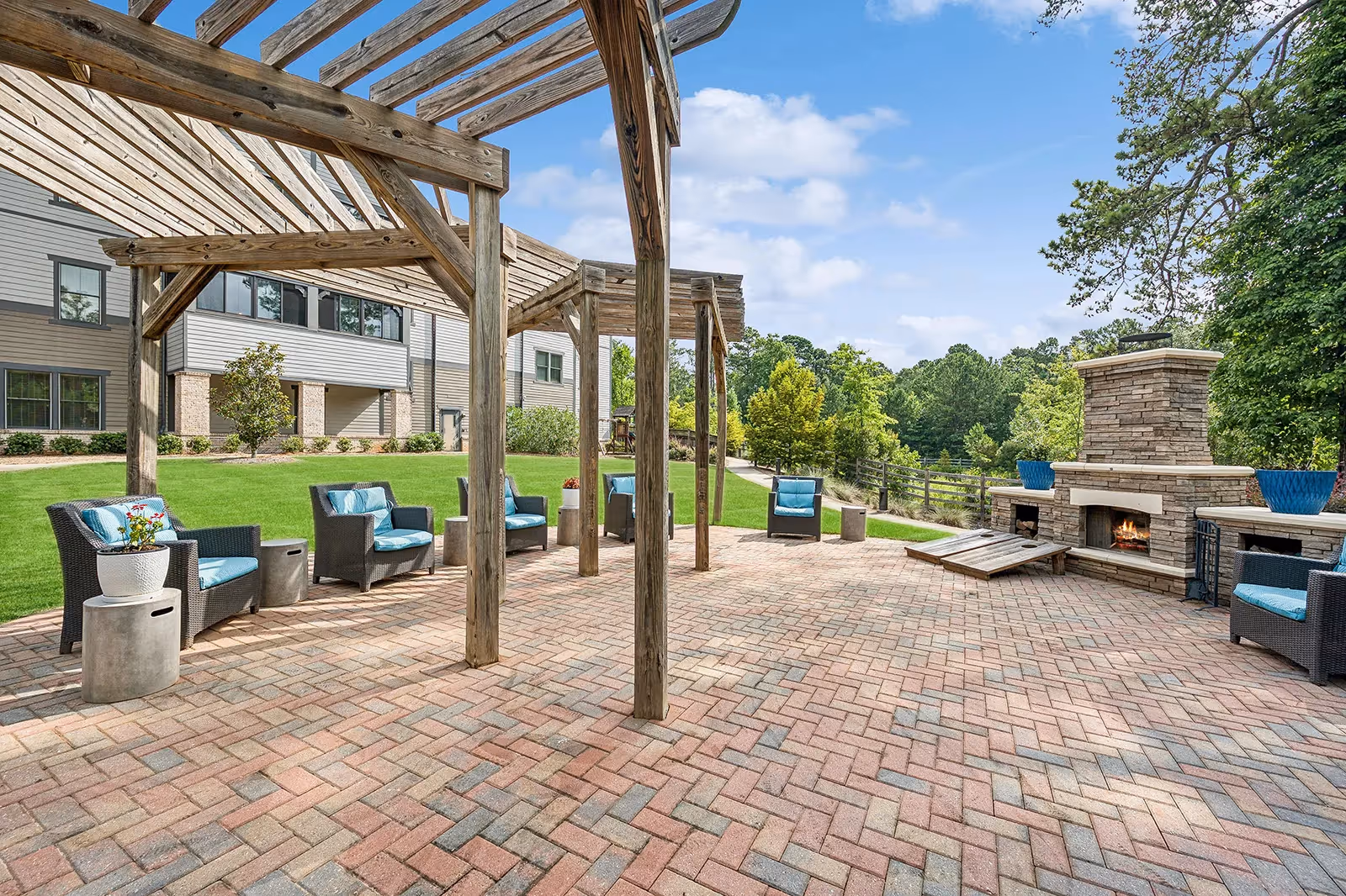 Outdoor patio with a wooden pergola, cushioned seating on brick pavers and a stone fireplace overlooking lawns and trees.