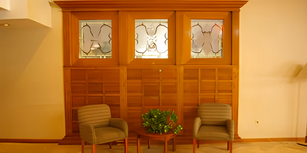 Two upholstered armchairs with striped fabric are positioned on either side of a small wooden table with a green leafy plant on it. Behind the chairs is a wooden panel wall with three decorative frosted glass windows featuring abstract designs. The room has beige walls and carpeted flooring.