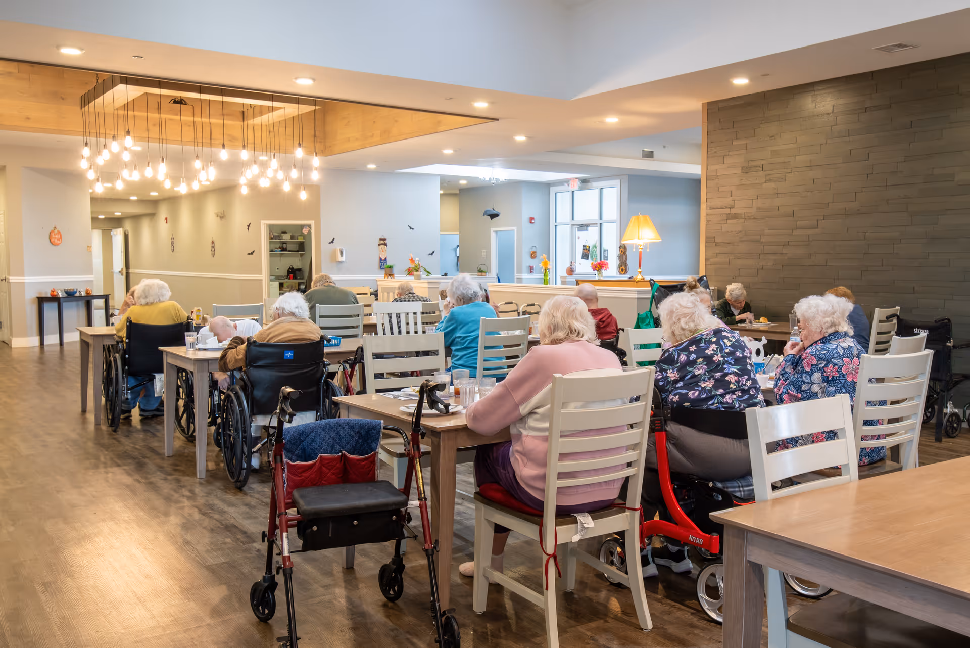 A group of elderly people sitting at tables in a well-lit dining area of a senior living facility. Some individuals are in wheelchairs, and others are seated on chairs. The room has wooden floors, modern hanging lights, and a cozy atmosphere with a lamp and decorations on a counter in the background.