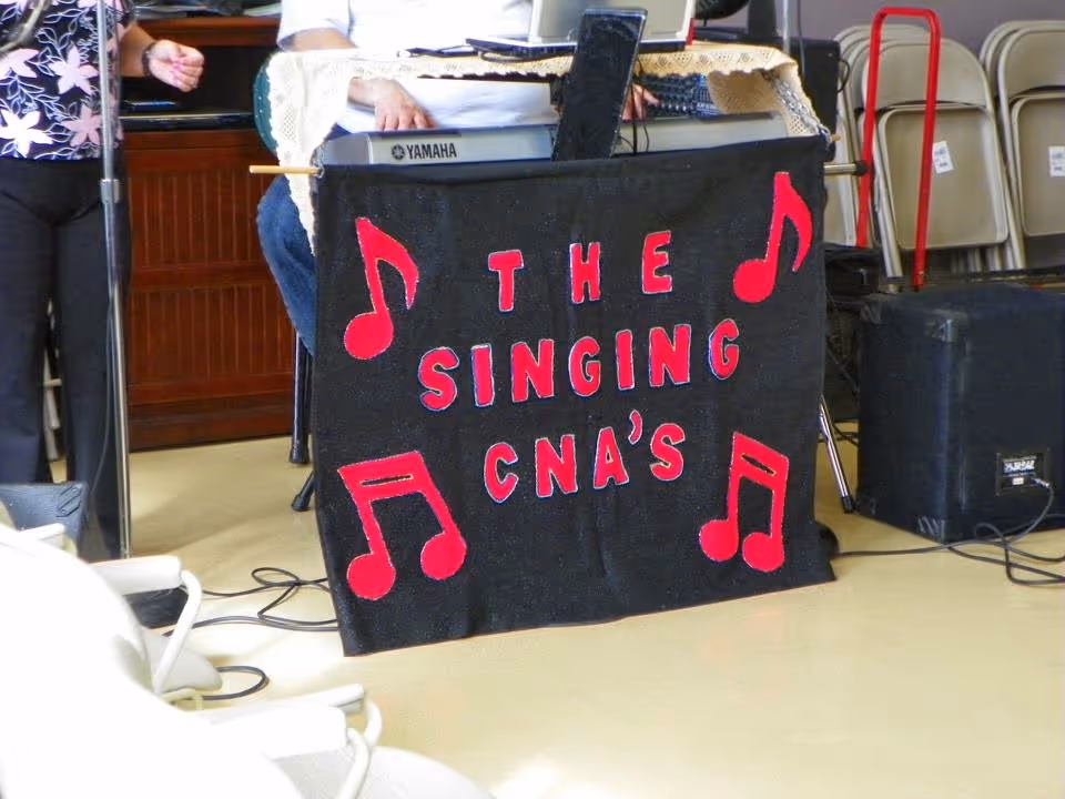 A keyboard draped with a black banner reading "THE SINGING CNA'S" with red musical notes, performers' hands, chairs and audio equipment in a community room.