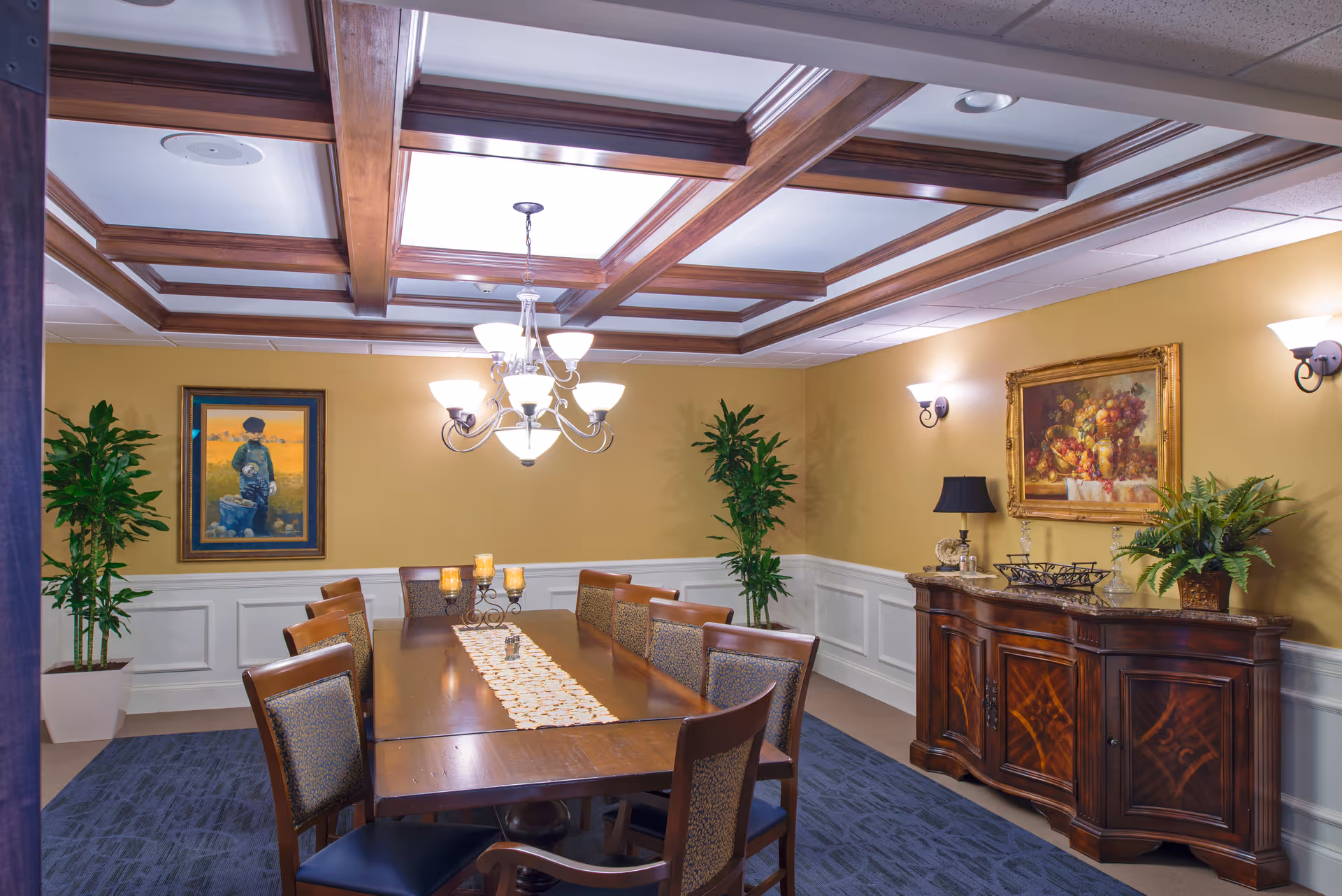 A formal dining room with a long wooden table surrounded by eight chairs. The room features a coffered ceiling with wooden beams and a chandelier hanging above the table. There are two potted plants in the corners, framed paintings on the walls, and a wooden sideboard with decorative items and a lamp. The walls are painted yellow with white wainscoting, and the floor is carpeted in a dark blue pattern.