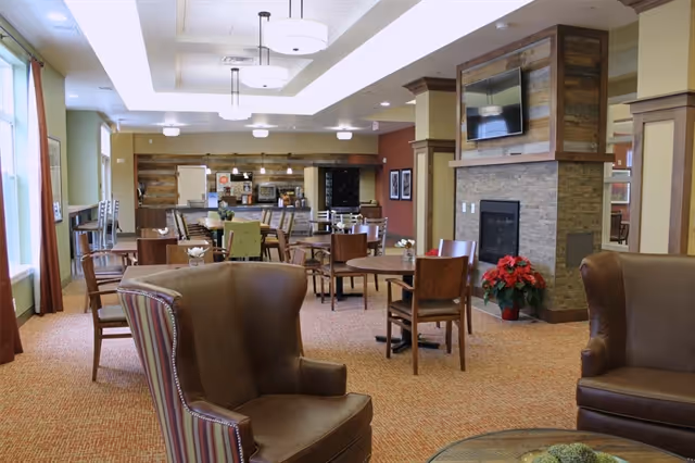 Spacious senior living common area with armchairs, dining tables, a fireplace with a TV, and a serving counter in the back.