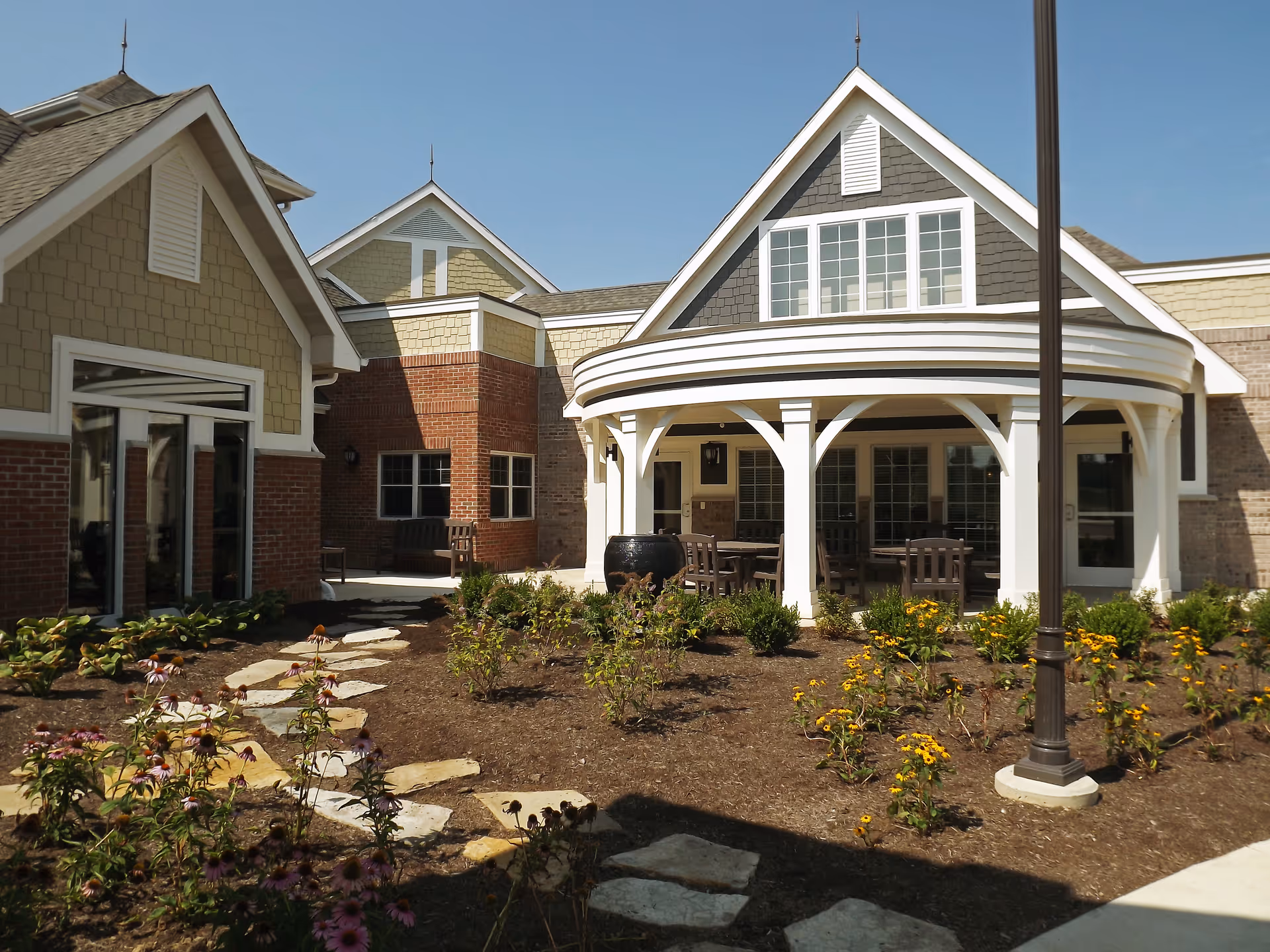 Exterior courtyard and entrance of a senior living building with a covered porch, outdoor seating, flower beds, and a stone pathway.