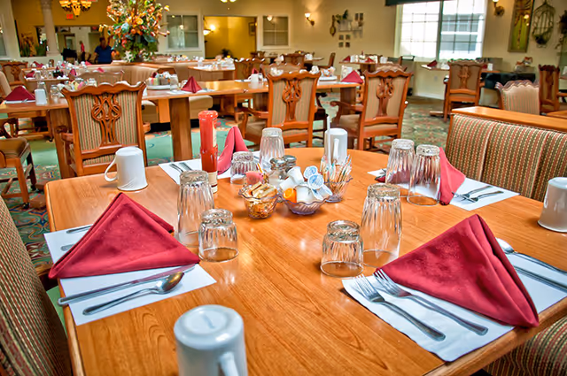 A dining room in an assisted living facility with wooden tables set for meals. Each table has red folded napkins, glasses turned upside down, white mugs, silverware, and condiments including ketchup and salt and pepper shakers. The room has wooden chairs with cushioned seats and a carpeted floor. Large windows and decorative plants are visible in the background.