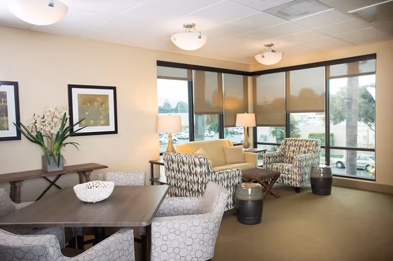 A bright and cozy living room area with large windows covered by beige roller shades. The room features a beige sofa, two patterned armchairs, a wooden coffee table, and two side tables with lamps. There is also a dining table with four upholstered chairs and a decorative bowl on top. A console table against the wall holds a vase with flowers and two framed artworks hang above it.