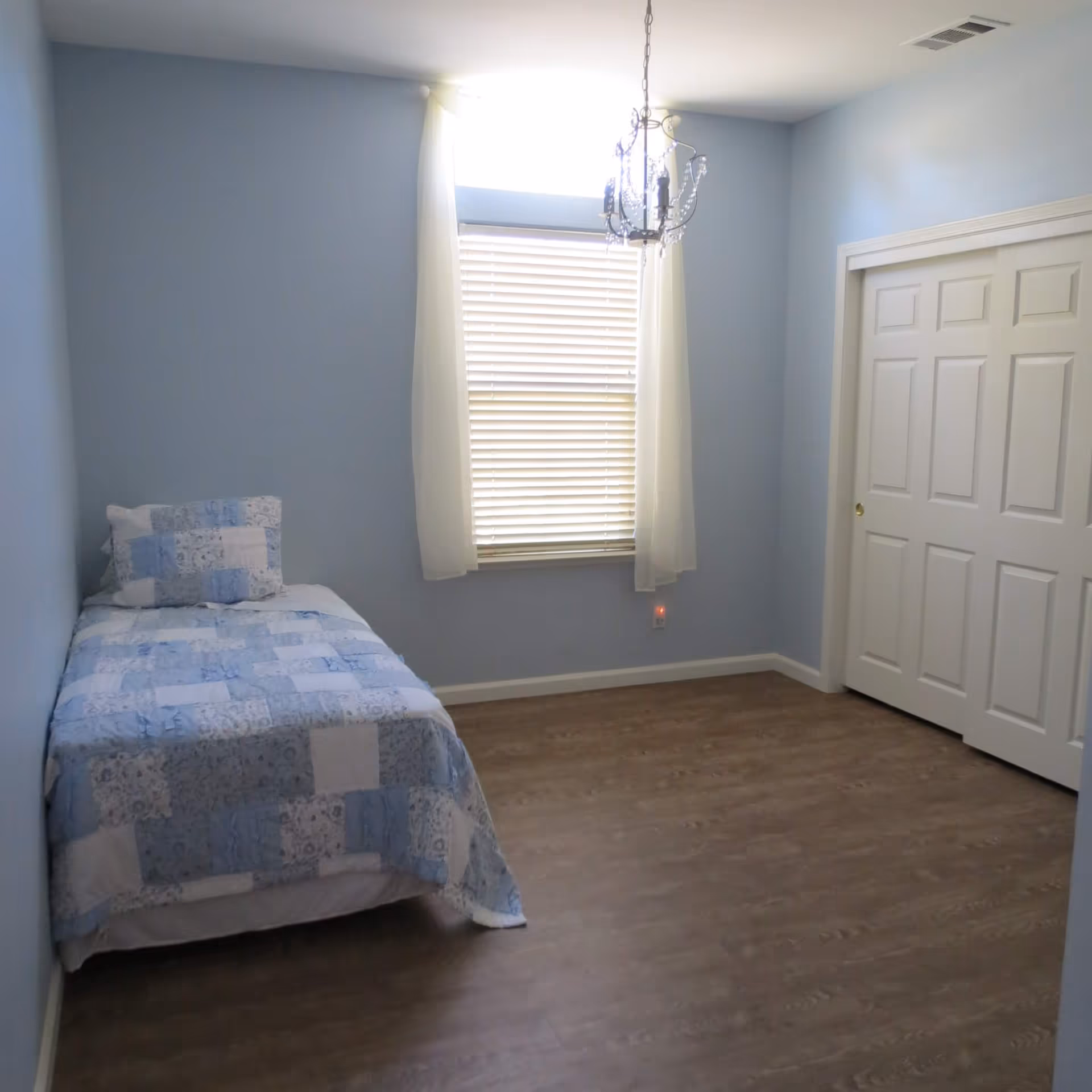 Small bedroom with a twin bed covered in a blue patchwork quilt, a window with blinds and white curtains, a hanging light fixture, and a closed closet.