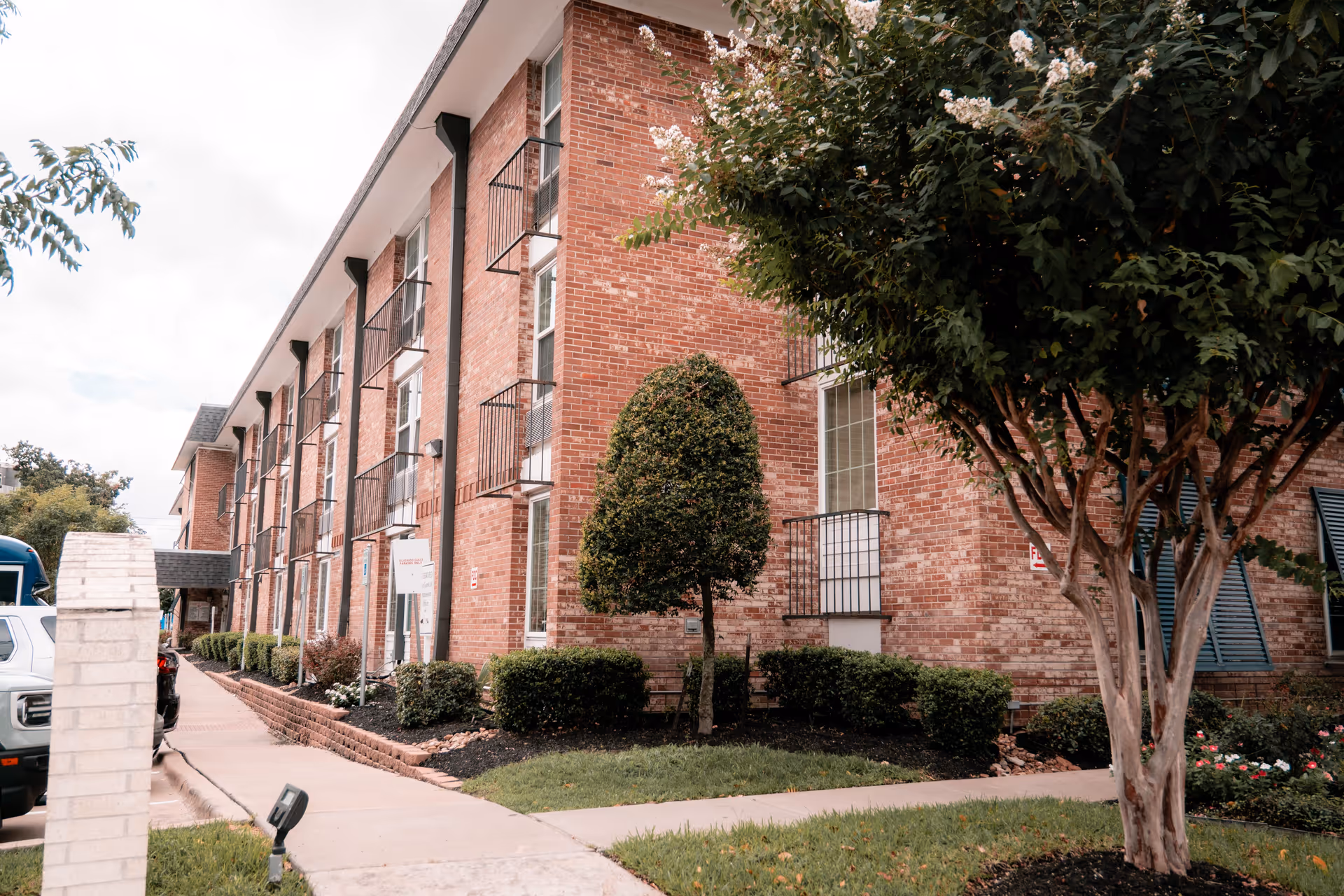 Red brick multi-story building with small balcony railings, a landscaped walkway and trees in front.