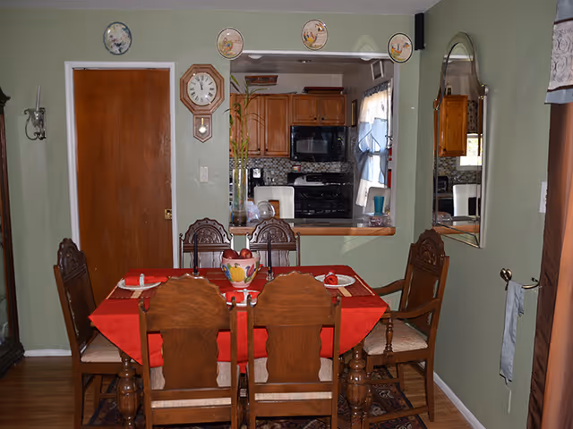 Dining room with a wooden table covered by a red tablecloth and six chairs, looking into a kitchen through a passthrough window.