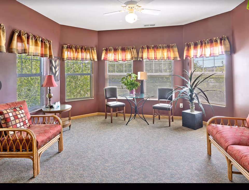 Sunlit common living area with wicker chairs and benches, a glass-top table, potted plants, and multiple bay windows.