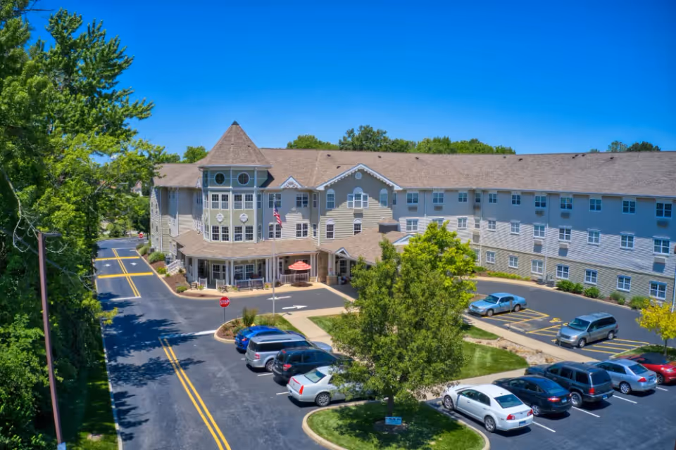 Exterior view of Addington Place of Collinsville, a large multi-story senior living facility with a peaked roof and multiple windows. The building is surrounded by a parking lot with several parked cars and landscaped greenery, including trees and bushes. The sky is clear and blue.