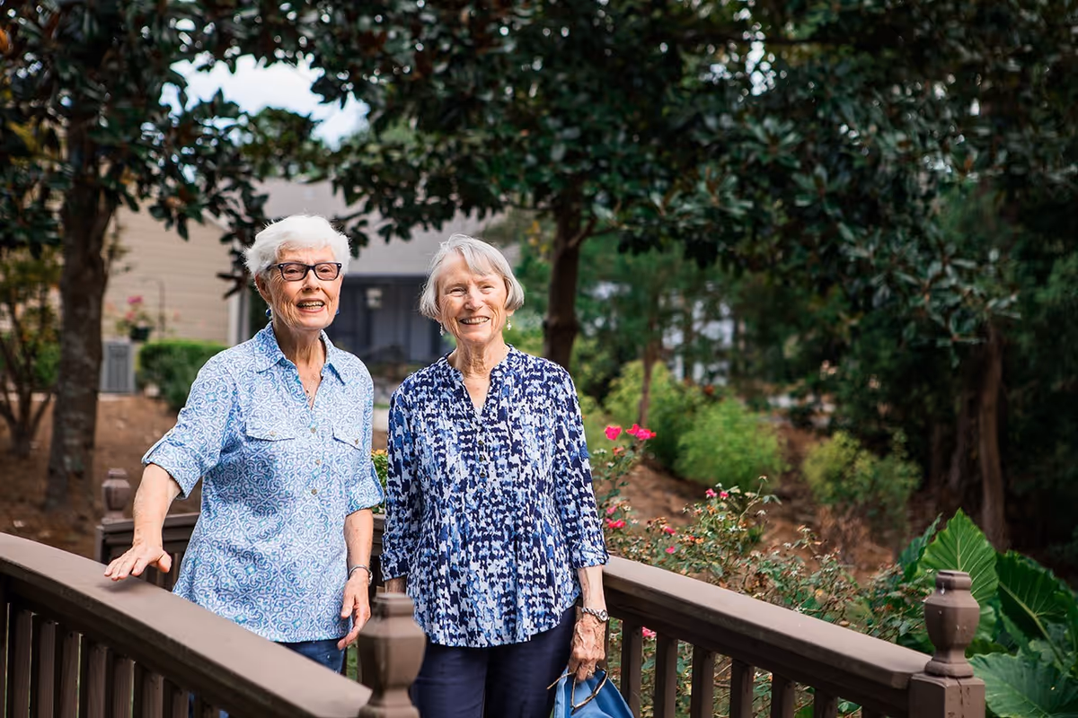 Two elderly women standing on a wooden bridge outdoors, smiling and enjoying a garden area with trees, flowers, and greenery in the background.