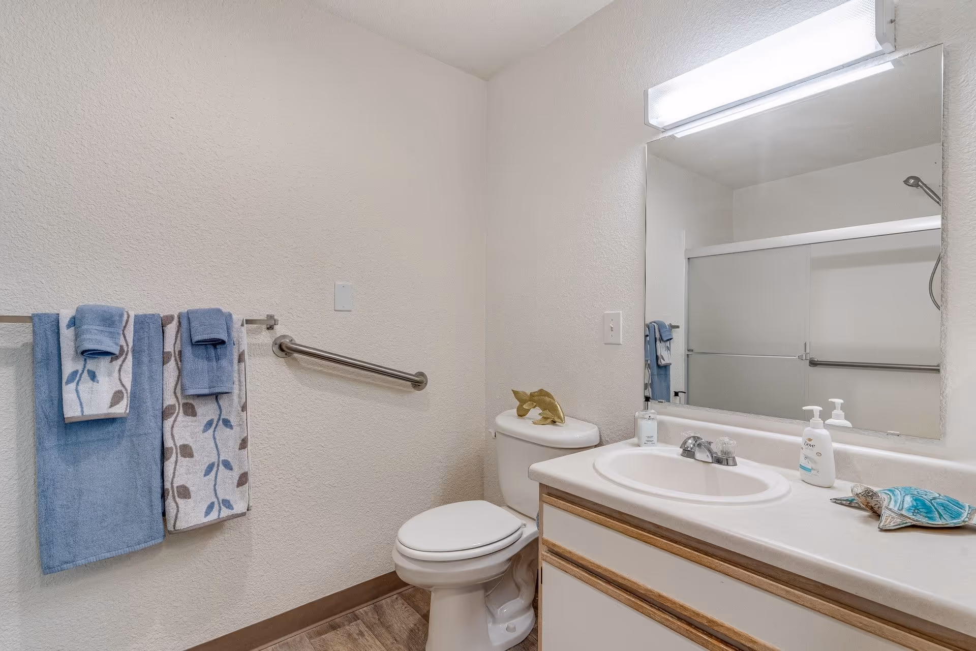 Small bathroom with a toilet, sink and mirror, blue towels on a rack and a sliding-glass shower door.